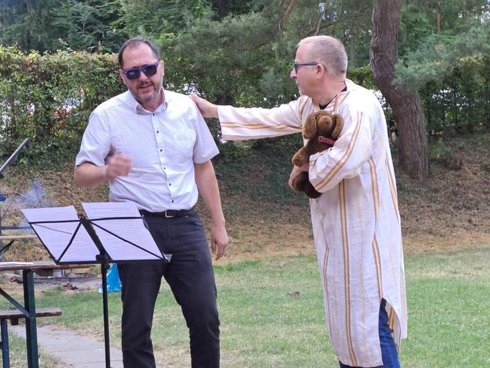 Sommerfest 2025 Begleitetes Wohnen in Familien (BWF) – Zwei Personen stehen an einem Notenständer, eine Person trägt einen Plüschhund in den Armen. 