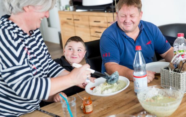 Drei Personen befinden sich an einem Tisch, dem sitzenden Kind in der Mitte wird von einer erwachsenen Person ein Teller gefüllt. Drei Personen befinden sich an einem Tisch, dem sitzenden Kind in der Mitte wird von einer erwachsenen Person ein Teller gefüllt.