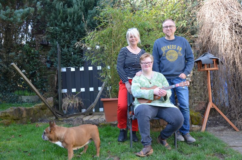 Vor Bäumen und Hecken stehen zwei Personen hinter einer dritten Person, die auf einem Stuhl sitzt und eine Gitarre in den Händen hält. Neben den drei Personen steht mit etwas Abstand ein Hund.