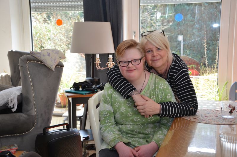 In einem Zimmer sitzt eine Person auf einem Stuhl. Eine zweite Person bückt sich zu ihr hinunter und legt ihre Arme um sie.